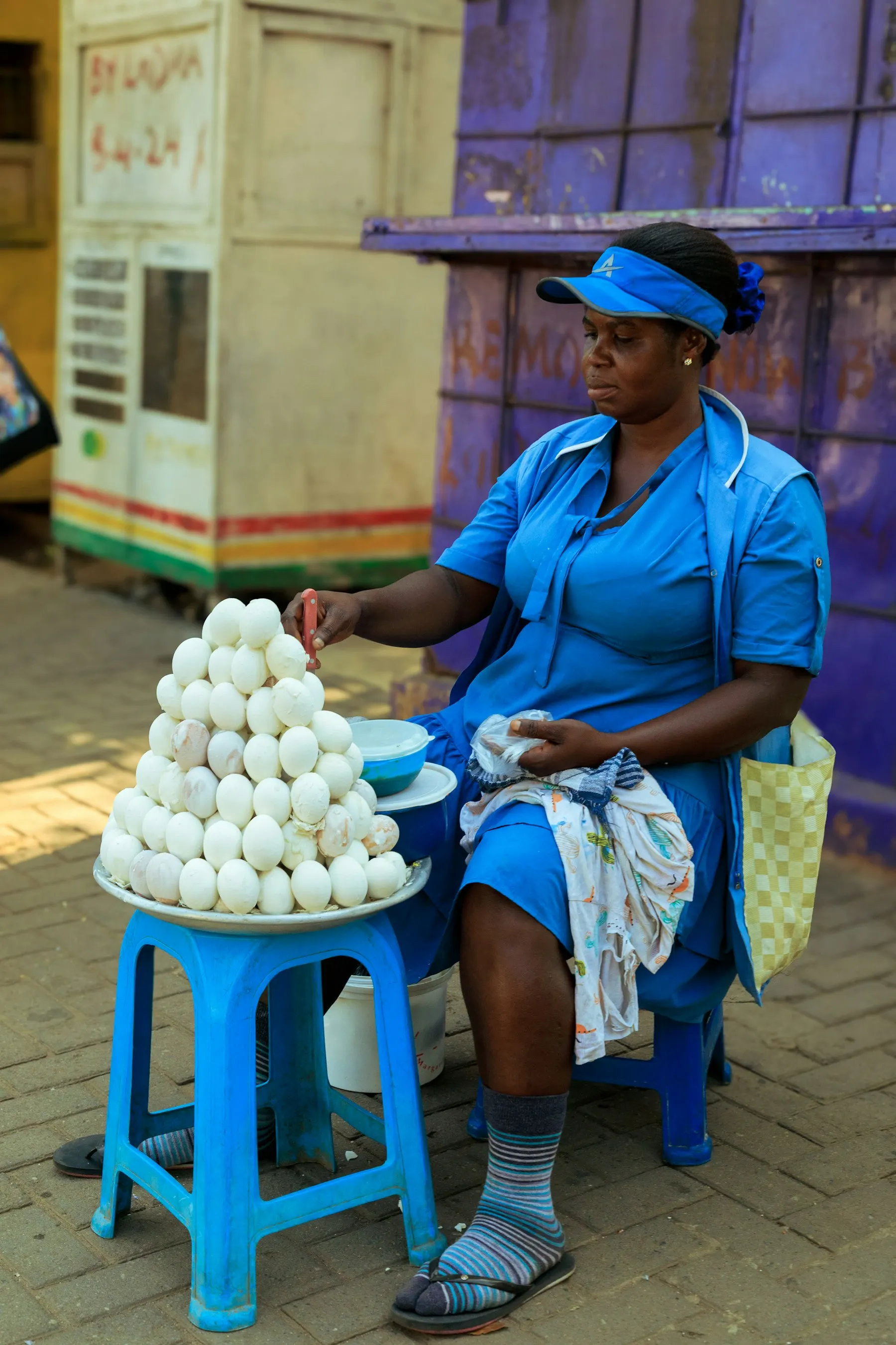 A woman in a blue uniform sits on a stool selling a pyramid of eggs