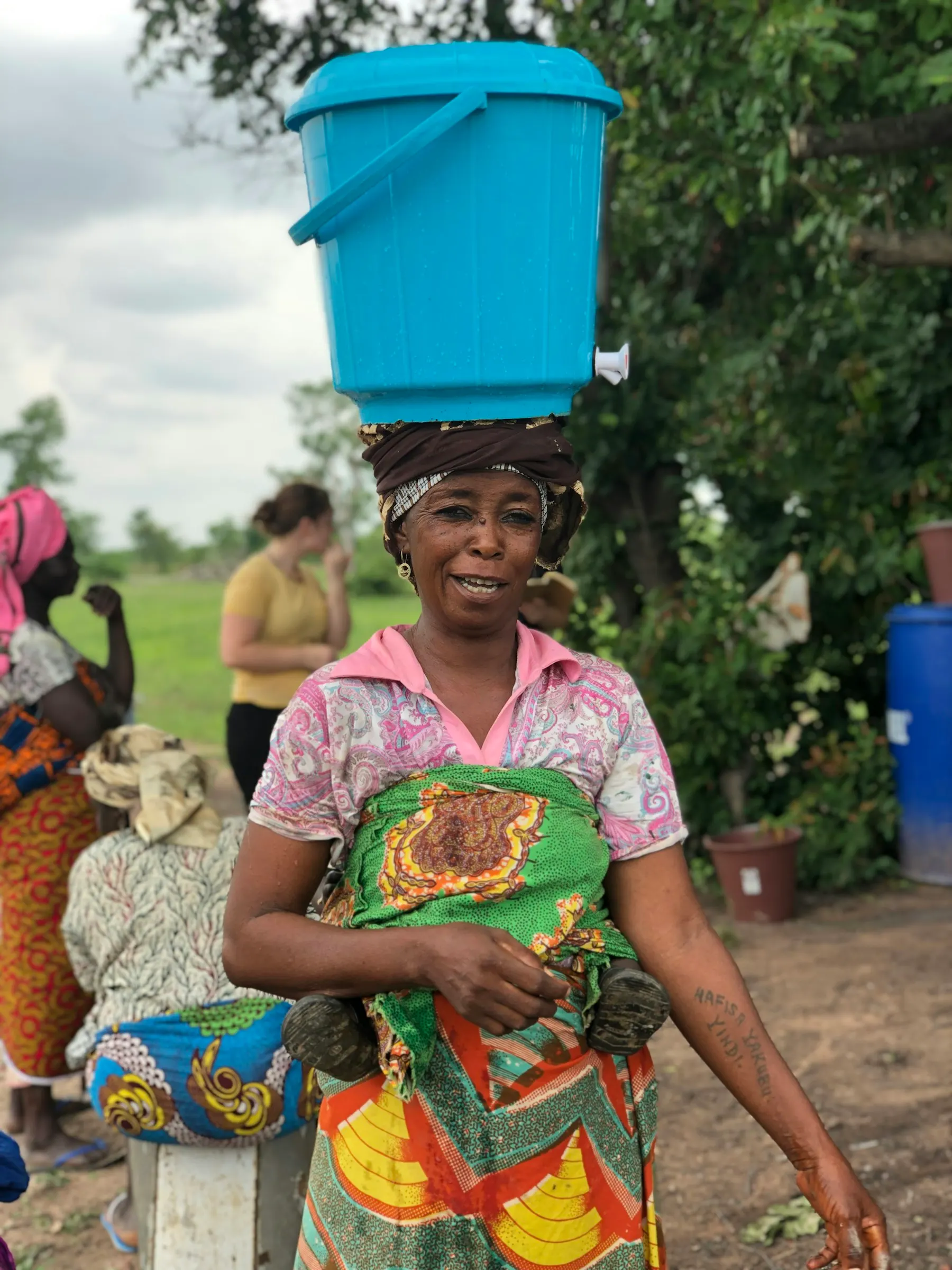 A smiling woman carrying a blue basin on her head, with a baby on her back